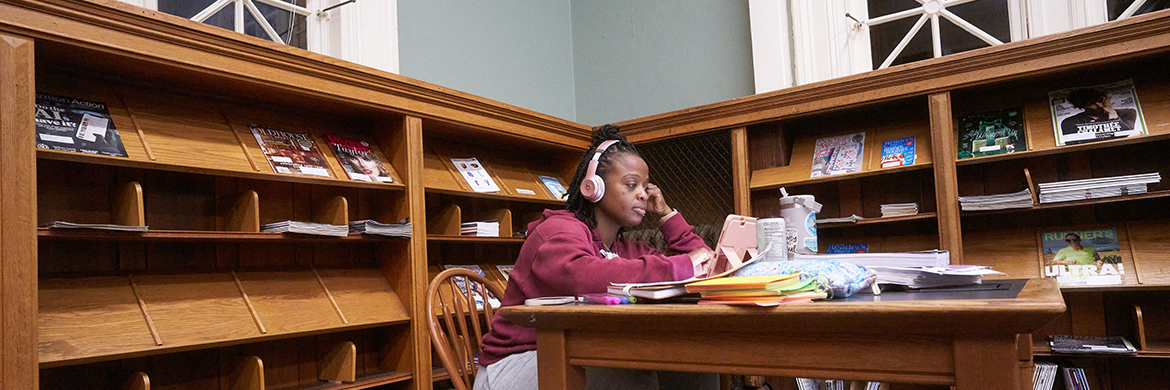 Woman with headphones on sitting at a table within the library with materials and a laptop on the desk