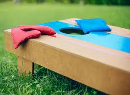 Green grass background with wooden cornhole board. There is a blue stripe on the cornhole board and 1 blue and 2 red bean bags sitting near the hole in the board.