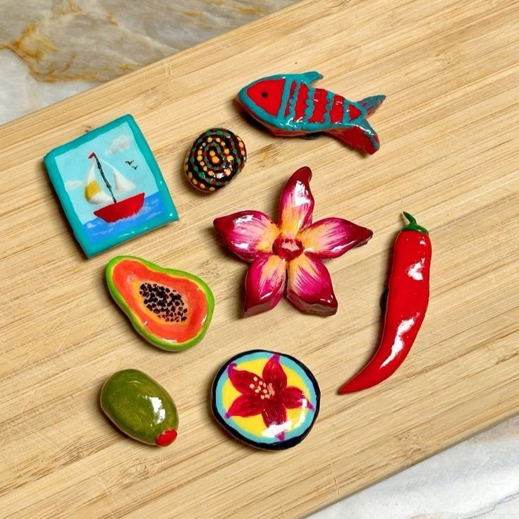 A collection of small clay magnets including a boat, fish, olive, papaya, pepper, and two flowers.